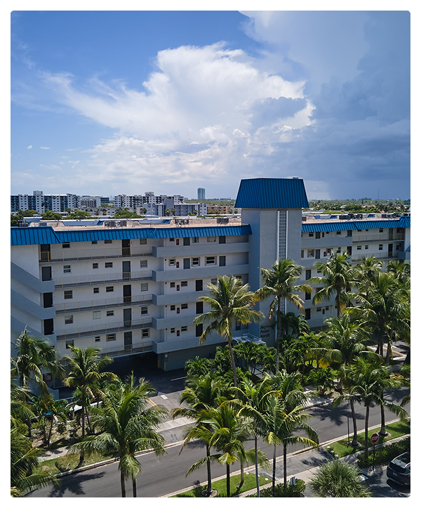 A multi-story white building with blue roof accents showcases expert commercial roofing, is surrounded by palm trees, and set against a cityscape with a cloudy sky. The weather appears sunny with some clouds.