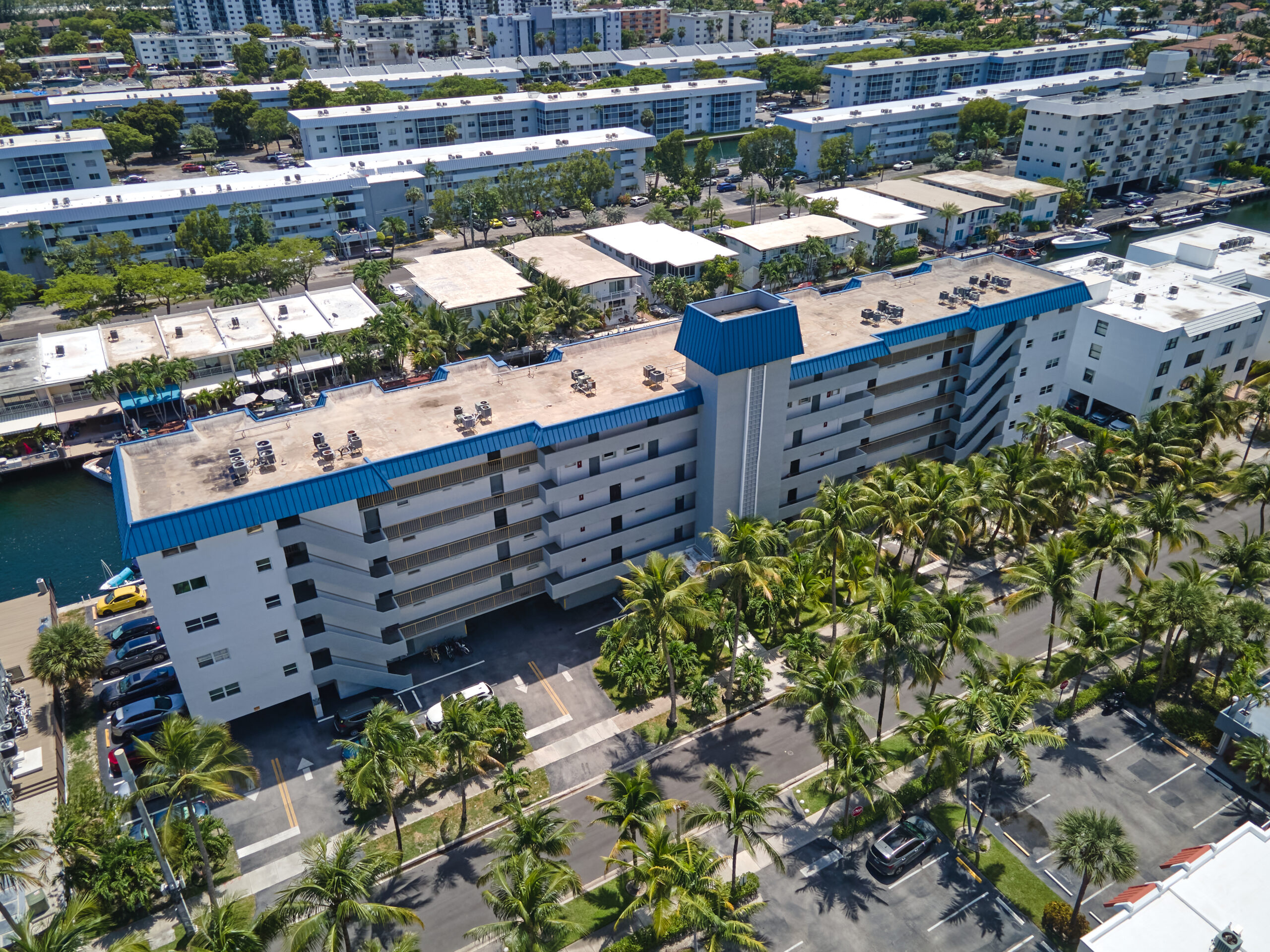 Aerial view of a large apartment building with a blue roofing in Miami, surrounded by palm trees and parking lots, with additional residential buildings and greenery in the background.