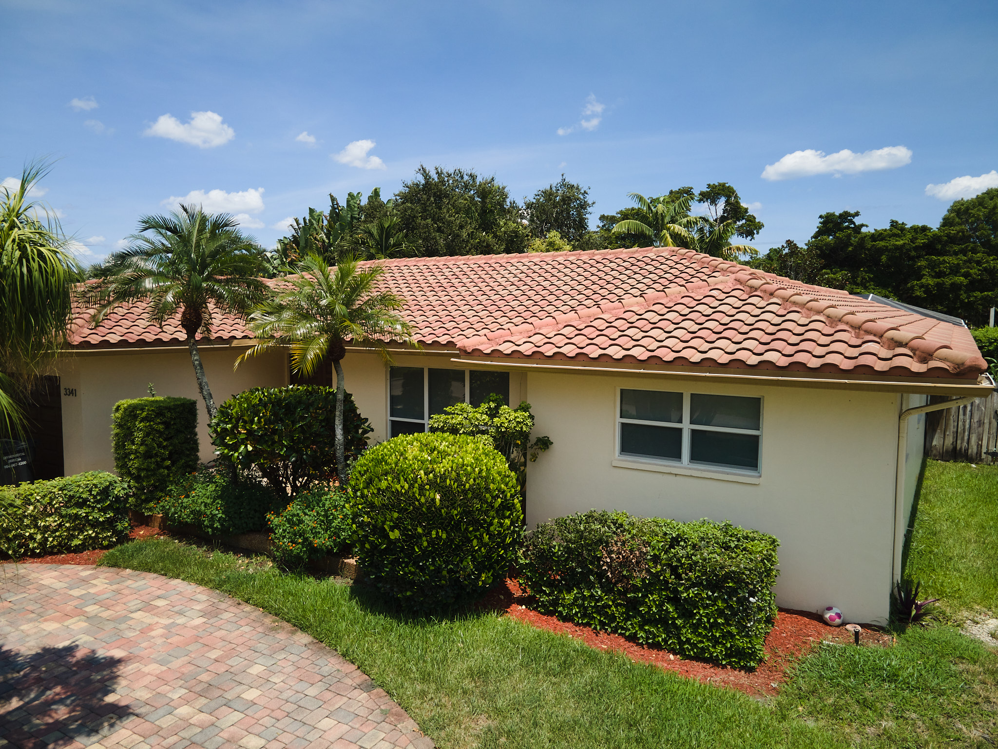 A single-story beige house in Miami with a red tile roofing, two windows, and neatly trimmed bushes and palm trees in the front yard, set under a blue sky with scattered clouds.